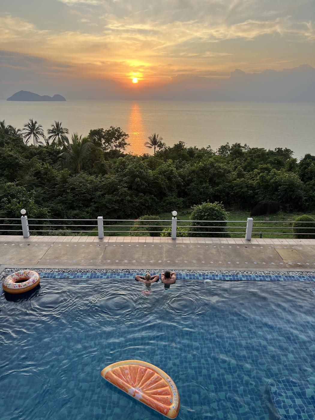 Two guests in the pool watching the sun set over the sea and a small island